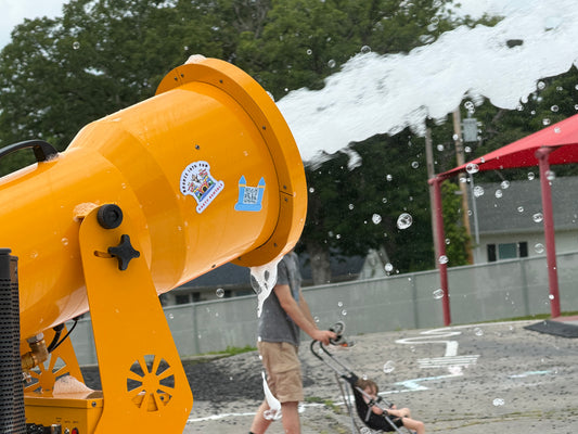 Orange snow blower with foam being dispensed, person operating it, and a child in a stroller nearby.